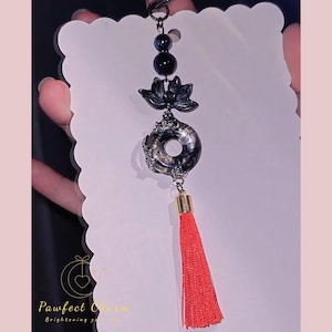 May include: A decorative charm featuring a black lotus flower, a circular pendant with a marbled effect, and a red tassel. The charm is suspended from a black chain with black beads. The charm is held in front of a white card.