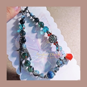 May include: A beaded keychain with a variety of colorful beads, including blue, green, and black. The keychain features a decorative sword charm and a red tassel. The text "Perfect Charm" is visible on the background.