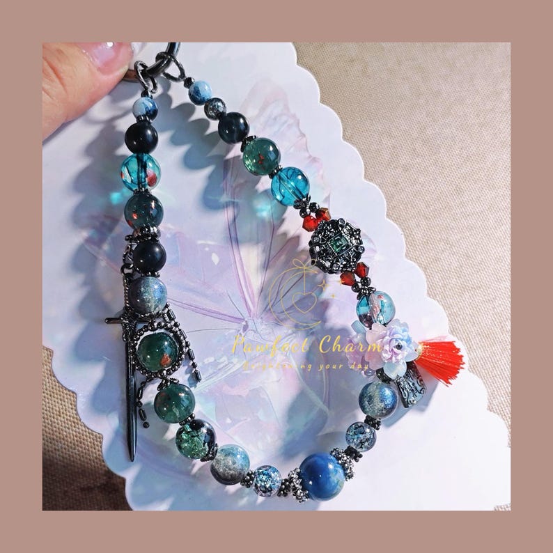 May include: A beaded keychain with a variety of colorful beads, including blue, green, and black. The keychain features a decorative sword charm and a red tassel. The text "Perfect Charm" is visible on the background.