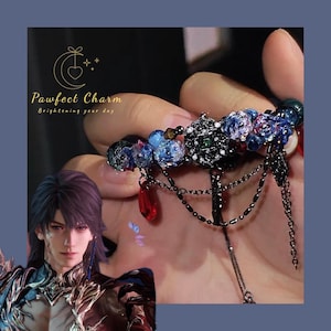 May include: An ornate, dark metal nail accessory with blue floral accents, red teardrop charms, and dangling chains. The accessory is held up by a hand. The image also includes the text "Pawfect Charm" and a graphic of a person.