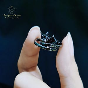 May include: A black ring with a detailed design featuring a crown and a dark gemstone. The ring is held between two fingers.