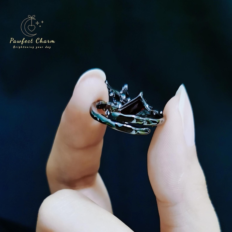 May include: A black ring with a detailed design featuring a crown and a dark gemstone. The ring is held between two fingers.