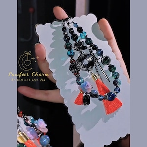 May include: A beaded charm bracelet with a variety of beads in shades of blue, green, and black. The charm includes a small sword, a red tassel, and a floral design. The text "Pawfect Charm Brightening your day" is visible.