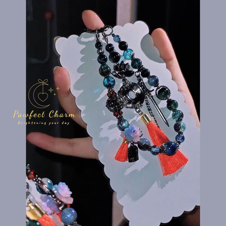 May include: A beaded charm bracelet with a variety of beads in shades of blue, green, and black. The charm includes a small sword, a red tassel, and a floral design. The text "Pawfect Charm Brightening your day" is visible.