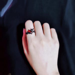 May include: A black ring with a red heart-shaped stone and intricate details. The ring is worn on a person's finger.