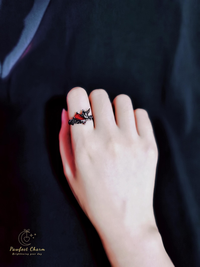 May include: A black ring with a red heart-shaped stone and intricate details. The ring is worn on a person's finger.