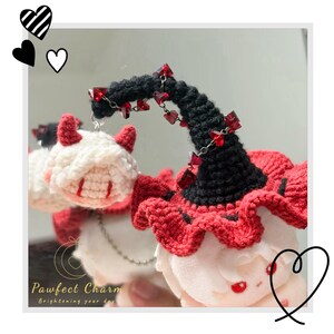 May include: A crochet plush doll featuring white hair, small red horns, and red eyes. The doll wears a black and red ruffled hat adorned with a silver chain and small red heart-shaped beads. The text Pawfect Charm Brightening your day is visible.