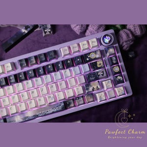 May include: A custom mechanical keyboard with a lavender case and white keycaps. The keycaps feature various designs, including celestial and floral motifs. The keyboard has a unique design with a light-up feature and the text "Pawfect Charm".