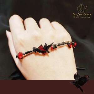 Op de afbeelding: Een zwarte armband met rode glazen kralen en een zwarte metalen bedel van twee vogels in vlucht. De bedel heeft de vorm van een hart. De armband zit om de pols van een persoon. De tekst "Pawfect Charm brightening your day" staat in de rechterbovenhoek van de afbeelding.