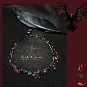 Op de afbeelding: Een zilveren ketting enkelbandje met rode glazen kralen en een bedel van een zwarte vogel met uitgespreide vleugels. Het enkelbandje ligt op een zwart oppervlak met een veer en de tekst "Pawfect Charm Brightening your day".
