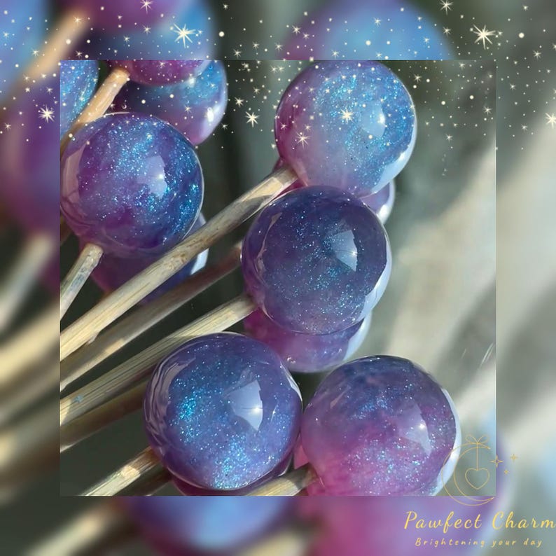 May include: Close-up of several round, galaxy-themed lollipops on wooden sticks. The lollipops have a gradient of purple and blue with glittery accents. The text "Pawfect Charm Brightening your day" is in the bottom right corner.