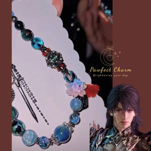 May include: A beaded bracelet with blue, green, and clear beads, a decorative centerpiece, and a small sword charm. The bracelet has a floral accent and a red tassel. The text "Pawfect Charm" is also visible.