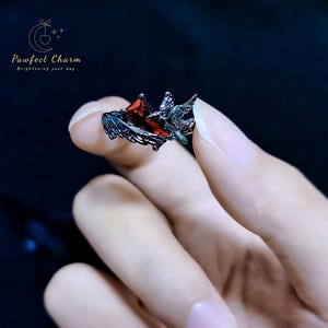 May include: A black ring with a red gemstone and a bird design. The ring is on a finger.