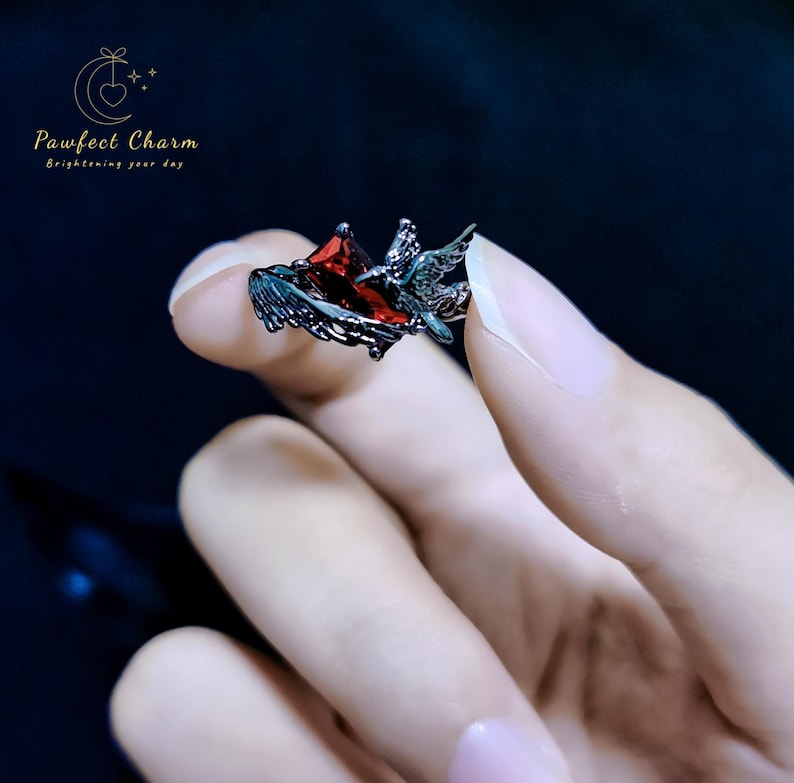 May include: A black ring with a red gemstone and a bird design. The ring is on a finger.