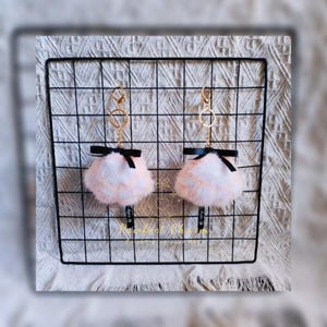 May include: Two fluffy keychains with a pink and white color scheme, each featuring a black bow and a gold-colored ring. The keychains are displayed on a black grid against a textured white background. The text "Pawfect Charm" is visible.