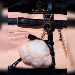 May include: A pink and black tote bag with a gold-colored chain and a fluffy, pink and white pom-pom keychain. The bag has a black strap with a silver buckle and a zipper. The text "Pawfect Charm" is visible.