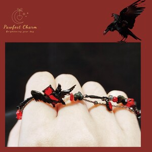 Op de afbeelding: Zwarte raven charme armband met rode glazen kralen en een zwarte raven hanger. De armband is op een hand.