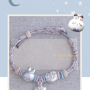 May include: A braided bracelet with pastel pink, blue, and gray threads. The bracelet features silver charms of a bunny, a boy, and a star with Chinese characters. The text "Pawfect Charm" is visible on the tag.