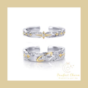 May include: Two silver rings with a gold accent. The top ring features a star design with a small clear stone in the center. The bottom ring features a planet design with a small clear stone in the center.