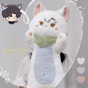 Op de afbeelding: Een grote witte pluche kat met roze oren, een groene sjaal en een lichtblauwe buik. De kat heeft geborduurde gezichtskenmerken en voetzolen. Een klein stripfiguur met kattenoren is zichtbaar.