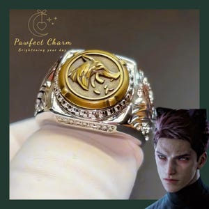 May include: A silver-toned ring featuring a circular gold-colored center inlay with a raised relief of a wolf head. The band has intricate side detailing and a row of small clear stones. The text Pawfect Charm Brightening your day is visible.