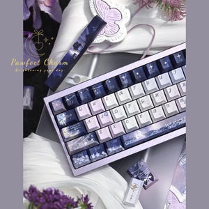 May include: A custom mechanical keyboard with a light purple frame and white keycaps. The keycaps feature a night sky design with stars and a landscape. The keyboard includes the text "Pawfect Charm Brightening your day".