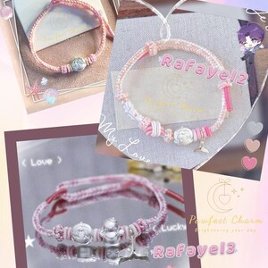 May include: Pink and white braided bracelets with silver charms. The bracelets have the text "Rafayel" and "Pawfect Charm". The image has a decorative background with hearts and a cartoon character.