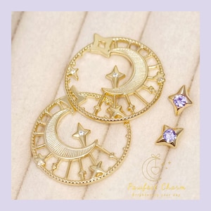 May include: Gold-colored celestial-themed jewelry set featuring two circular pendants with crescent moons, stars, and sun ray designs. Two star-shaped stud earrings with purple gemstones are also included. The jewelry is displayed on a light beige surface.