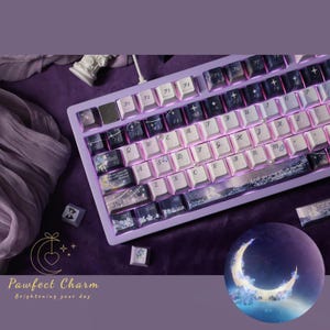 May include: A lavender mechanical keyboard with white and blue keycaps. The keycaps have a starry night design. The keyboard has a purple backlight. The text "Pawfect Charm Brightening your day" is in the lower left corner.