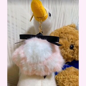 May include: A white plush duck keychain with a yellow beak and gold-colored hardware. It features a fluffy pink and white pom-pom with a black bow. A brown teddy bear is partially visible in the background.