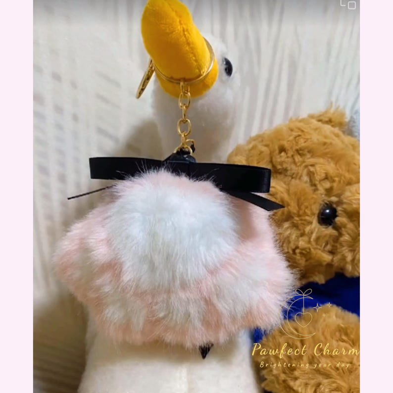 May include: A white plush duck keychain with a yellow beak and gold-colored hardware. It features a fluffy pink and white pom-pom with a black bow. A brown teddy bear is partially visible in the background.