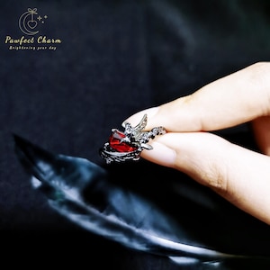 May include: A black ring with a red gemstone and a silver bird design. The ring is on a finger.