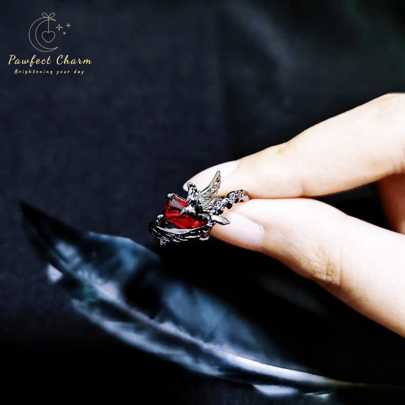 May include: A black ring with a red gemstone and a silver bird design. The ring is on a finger.
