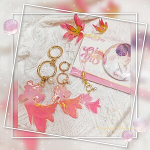 May include: Pink and gold keychains with a dragon design, alongside a pin featuring a person with purple hair. The keychains have gold rings and pearl accents. The pin is on a pink card with text.