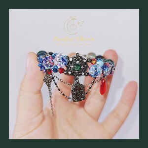 May include: A beaded bracelet with a central decorative element featuring a green gem and intricate metalwork. The bracelet incorporates blue floral beads, red accents, and dangling charms. The text "Fanfest Charm" is visible in the background.