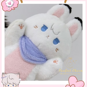 Op de afbeelding: Een pluche wit konijn met blauwe ogen, roze binnenoren en een lichtpaarse sjaal. Het konijn heeft een roze borst en pootkussentjes. De afbeelding bevat een cartoon kattenpersonage en de tekst "Pawfeel Charm".