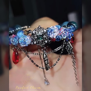 May include: A detailed bracelet featuring blue floral beads, dark metal charms, and dangling red teardrop-shaped beads. The bracelet has a central decorative element with a green gem. The text "Pawfect Charm" is visible.