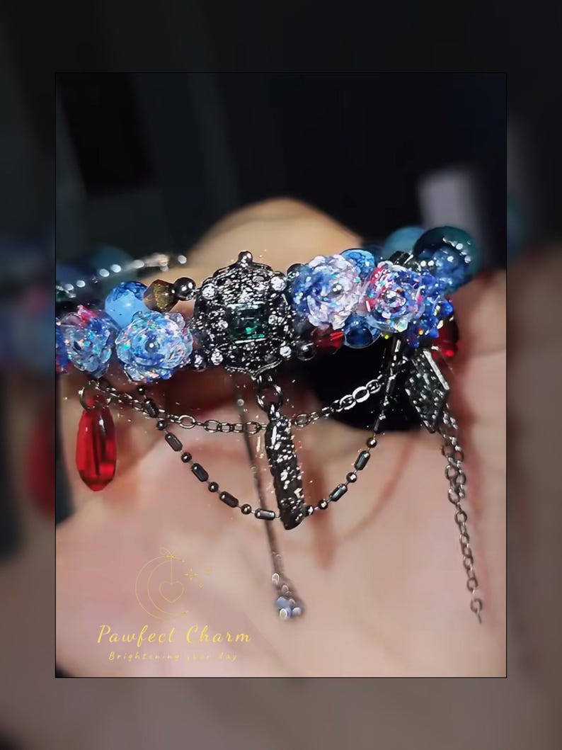 May include: A detailed bracelet featuring blue floral beads, dark metal charms, and dangling red teardrop-shaped beads. The bracelet has a central decorative element with a green gem. The text "Pawfect Charm" is visible.