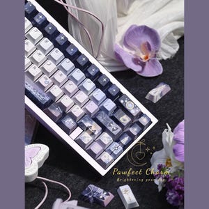 May include: A custom mechanical keyboard with keycaps in shades of purple, white, and blue. The keycaps feature various designs, including stars and celestial patterns. The keyboard is angled, and the text "Pawfect Charm Brightening your day" is visible.