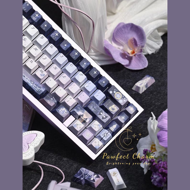 May include: A custom mechanical keyboard with keycaps in shades of purple, white, and blue. The keycaps feature various designs, including stars and celestial patterns. The keyboard is angled, and the text "Pawfect Charm Brightening your day" is visible.