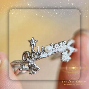 May include: A silver ring with a delicate branch design, featuring the word "Adore" and a small pearl. A star sits atop the branch.