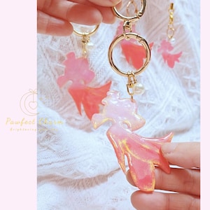 May include: Pink and gold resin keychains with a stylized fish design. The keychains have gold-colored hardware, including a large ring and a small pearl charm. The fish-shaped charms have a gradient of pink and gold colors.