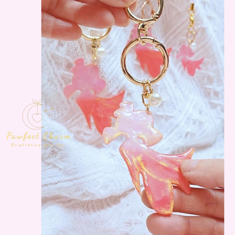 May include: Pink and gold resin keychains with a stylized fish design. The keychains have gold-colored hardware, including a large ring and a small pearl charm. The fish-shaped charms have a gradient of pink and gold colors.