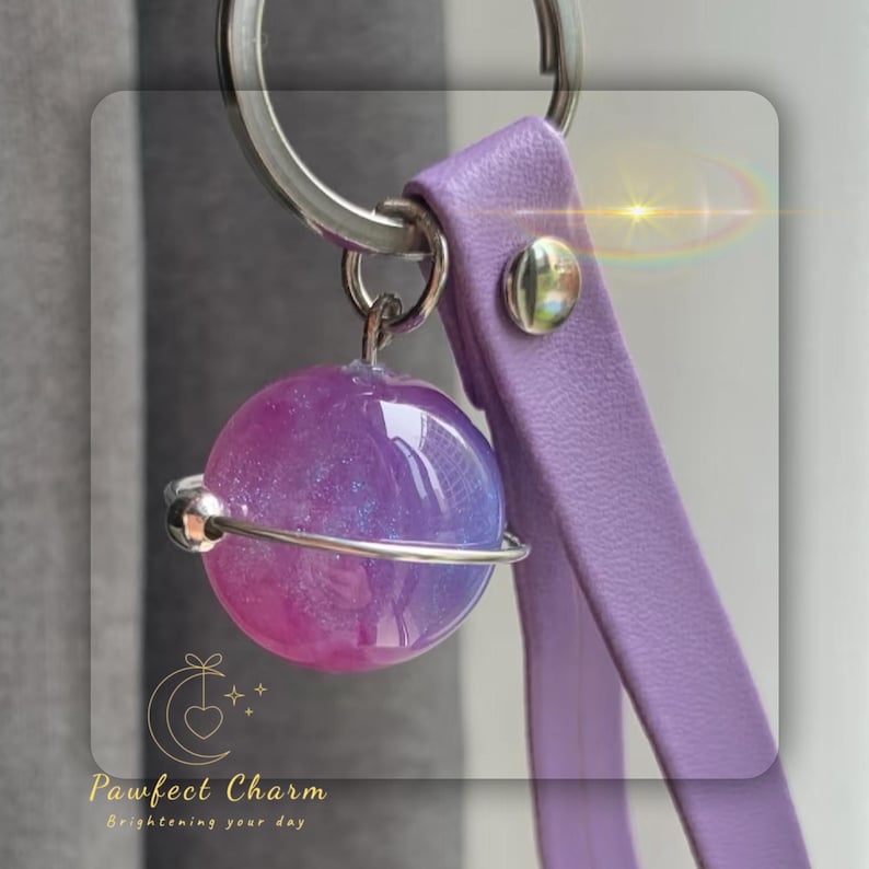 May include: A keychain featuring a purple and pink planet-shaped charm with a silver band. The charm is attached to a lavender leather strap and a silver keyring. The text "Pawfect Charm Brightening your day" is visible.