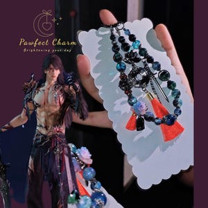 May include: A beaded charm bracelet with blue, black, and green beads, red tassels, and a silver sword charm. The bracelet is held against a white background with the text "Pawfect Charm Brightening your day".