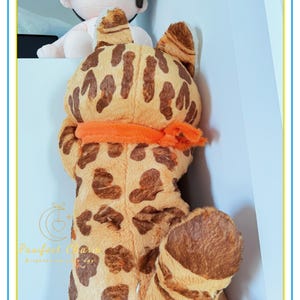 Op de afbeelding: Een groot pluche speelgoed in de vorm van een Bengaalse kat, met een bruin en beige gevlekt patroon. Het heeft grote oren, een oranje sjaal en een zacht, knuffelbaar uiterlijk. Op de achtergrond is een kleinere pop te zien.