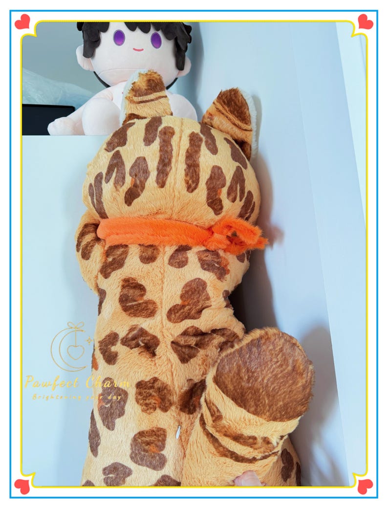 Op de afbeelding: Een groot pluche speelgoed in de vorm van een Bengaalse kat, met een bruin en beige gevlekt patroon. Het heeft grote oren, een oranje sjaal en een zacht, knuffelbaar uiterlijk. Op de achtergrond is een kleinere pop te zien.