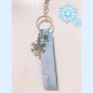 May include: A light blue leather keychain with a silver snowflake charm. The keychain has a silver ring and a snap closure. The background has a light blue watercolor effect and the text "Pawfect Charm".