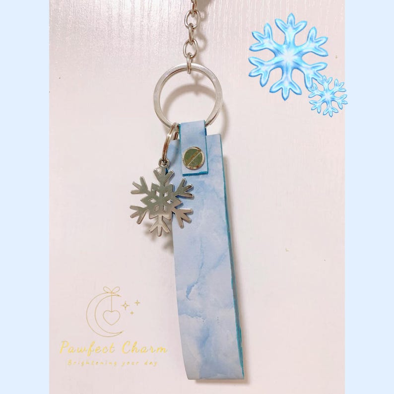 May include: A light blue leather keychain with a silver snowflake charm. The keychain has a silver ring and a snap closure. The background has a light blue watercolor effect and the text "Pawfect Charm".