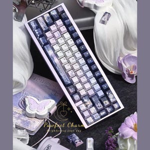 May include: A custom mechanical keyboard with a lavender and white color scheme. The keycaps feature a gradient design and various decorative icons. The keyboard is surrounded by accessories, including a butterfly-shaped item and a book.